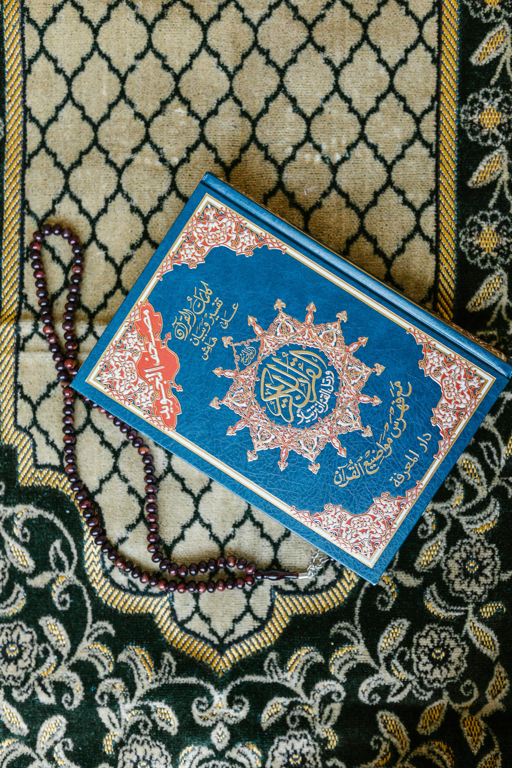 Top view of a Quran with prayer beads on a decorative Islamic carpet.